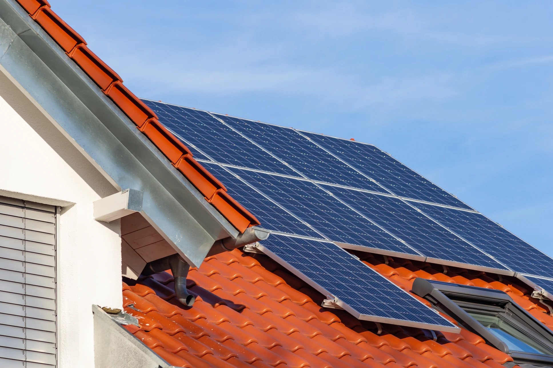Deux techniciens en combinaisons vertes et casques blancs installent des panneaux photovoltaïques sur une toiture résidentielle en tuiles sous un ciel bleu dégagé, environnement verdoyant visible en arrière-plan